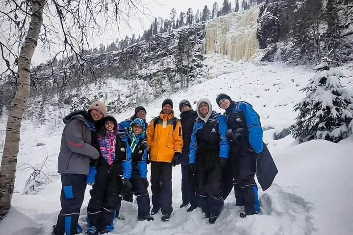 A tour group in thermal overalls standing in the snow below the frozen waterfalls of Korouoma Canyon